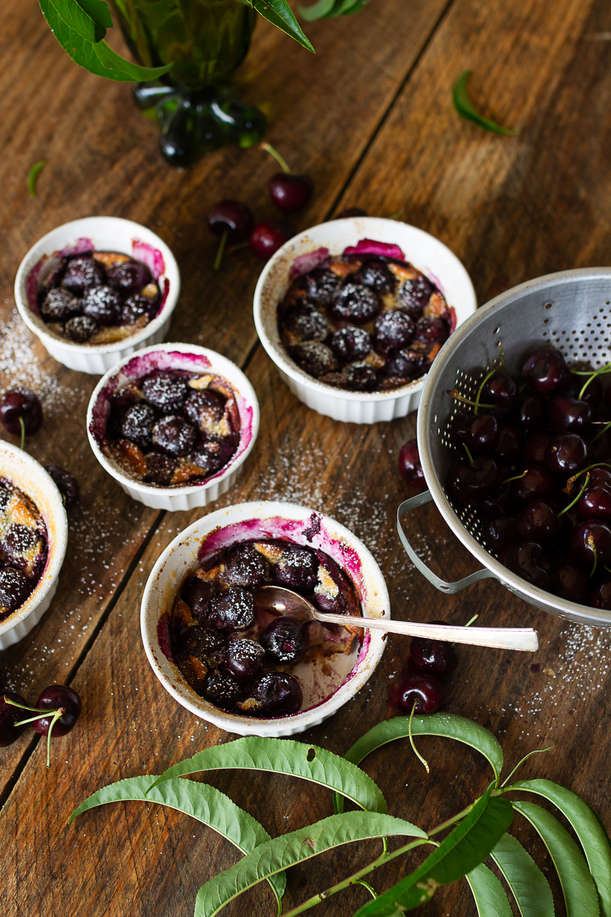 Rustic farmhouse table setting featuring four individual sweet cherry clafoutis baked in white ramekins with powdered sugar dusting and a metal colander of fresh fruit.