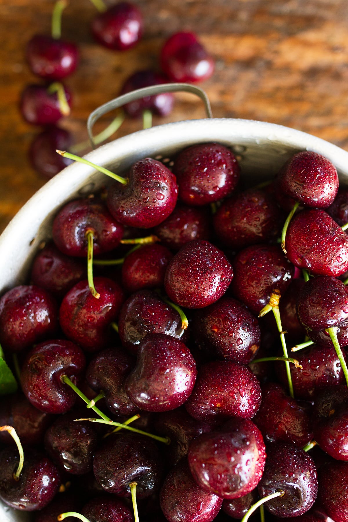 Macro shot focusing on glossy, ripe dark sweet cherries piled high in a rustic metal straining basket.