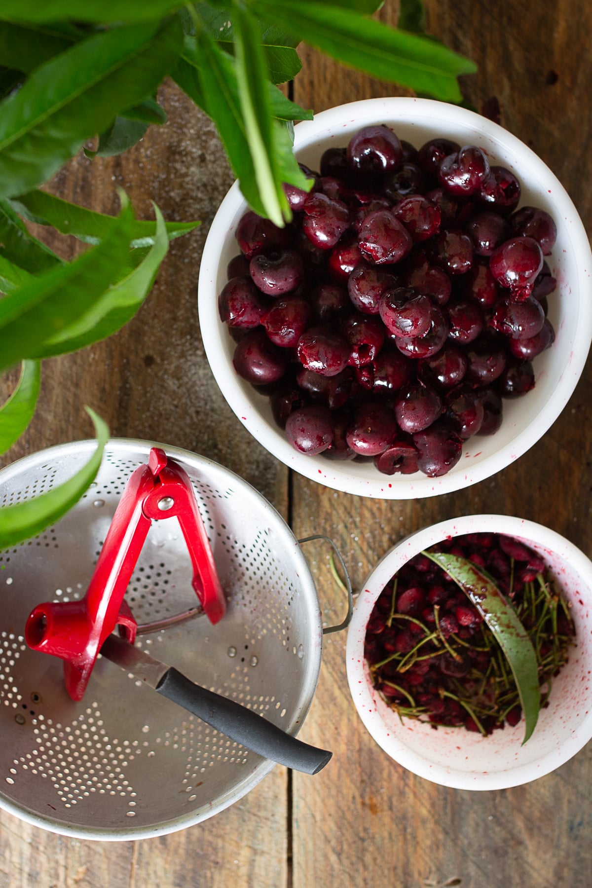 Overhead view of fresh cherries being prepared on a wooden surface with a red handheld cherry pitter, an empty colander, and a bowl filled with discarded cherry pits and stems.