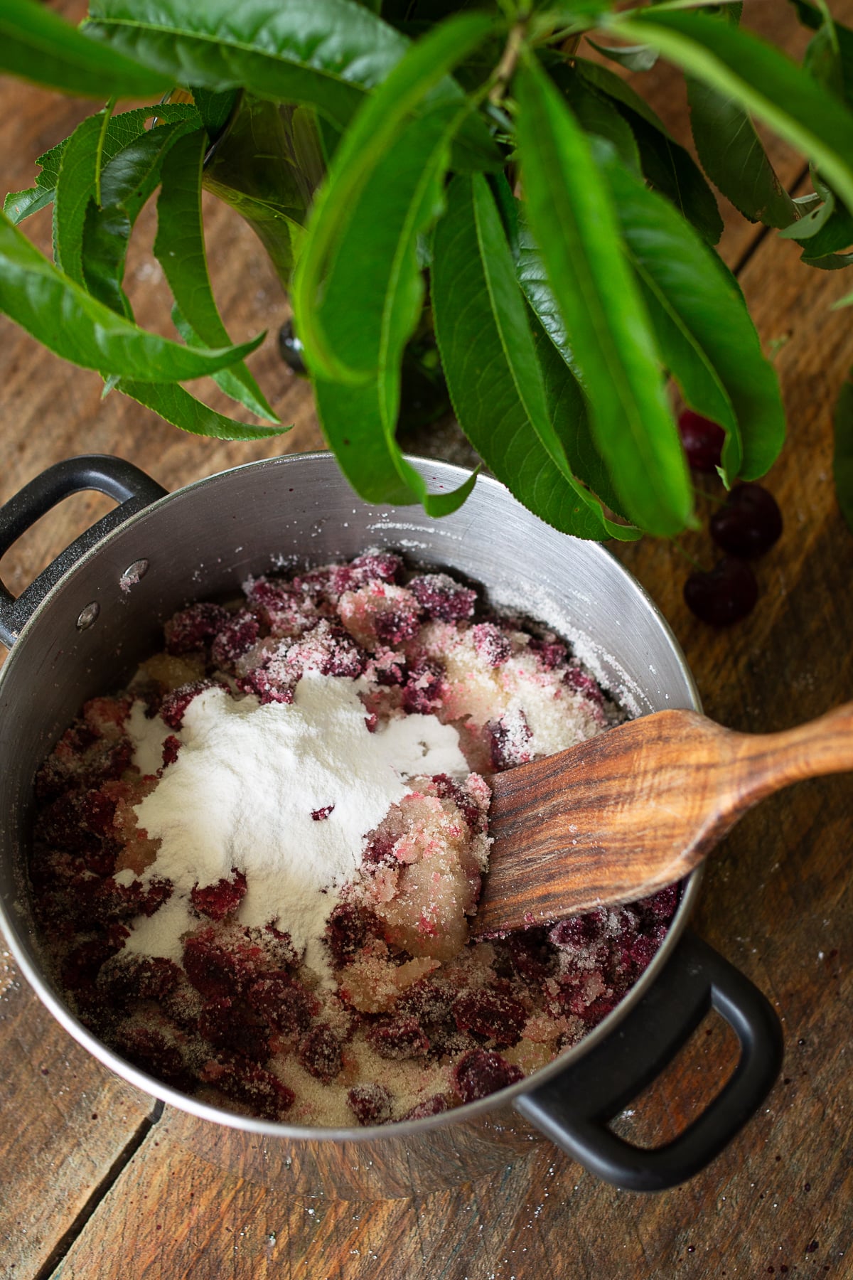 Sliced cherries mixed with granulated sugar and powdered pectin inside a stainless steel stockpot with a large wooden spatula.