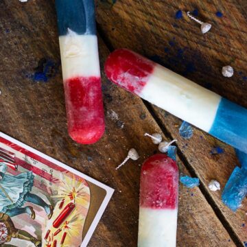 Three homemade rocket pops on a wooden background next to a vintage American flag postcard and small shards of blue ice.
