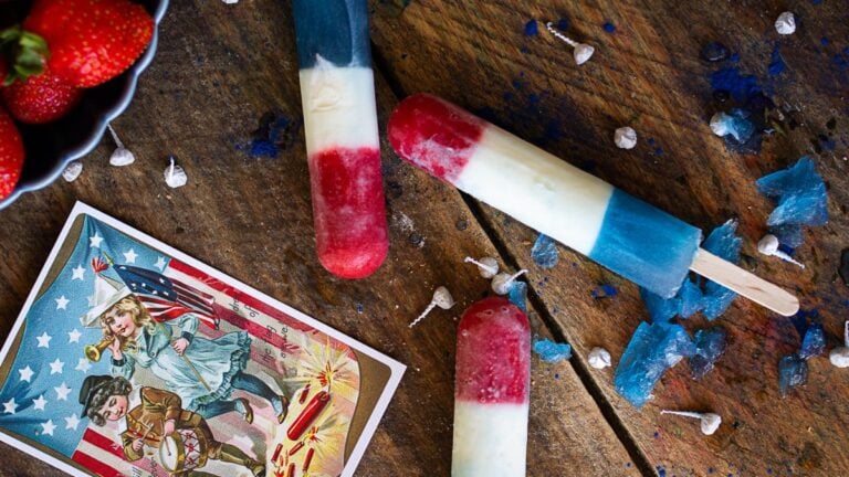 Three homemade rocket pops on a wooden background next to a vintage American flag postcard and small shards of blue ice.