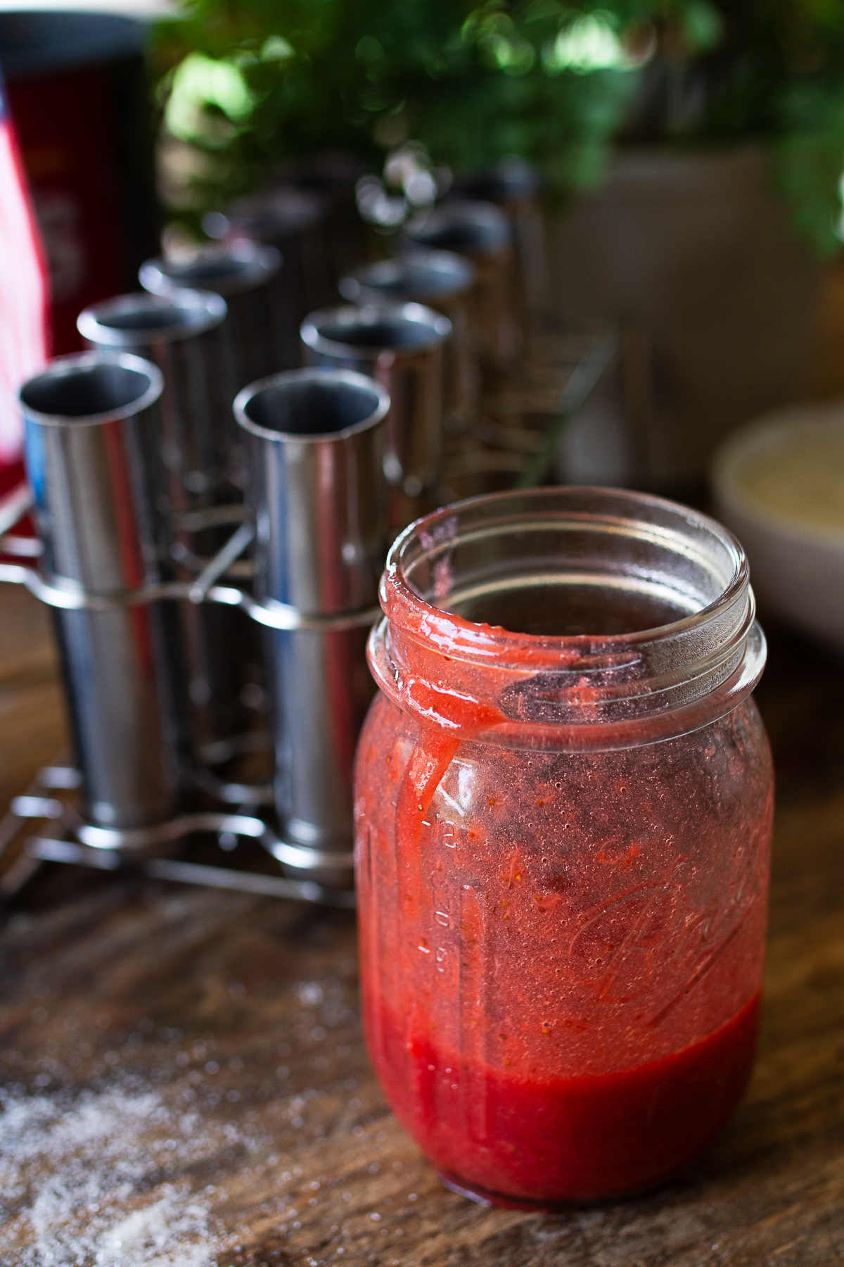 A clear glass mason jar filled with bright red strawberry puree on a rustic wooden table next to a stainless steel popsicle mold.
