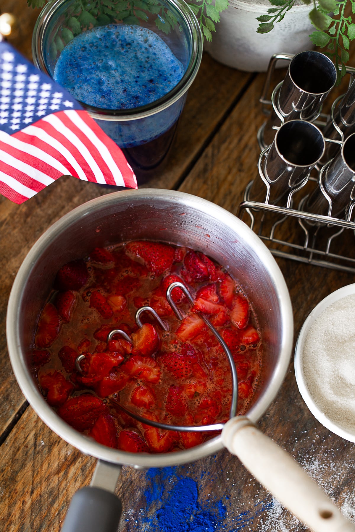 Overhead view of fresh strawberries being mashed in a stainless steel pot next to a glass of vibrant blue spirulina liquid and an American flag.
