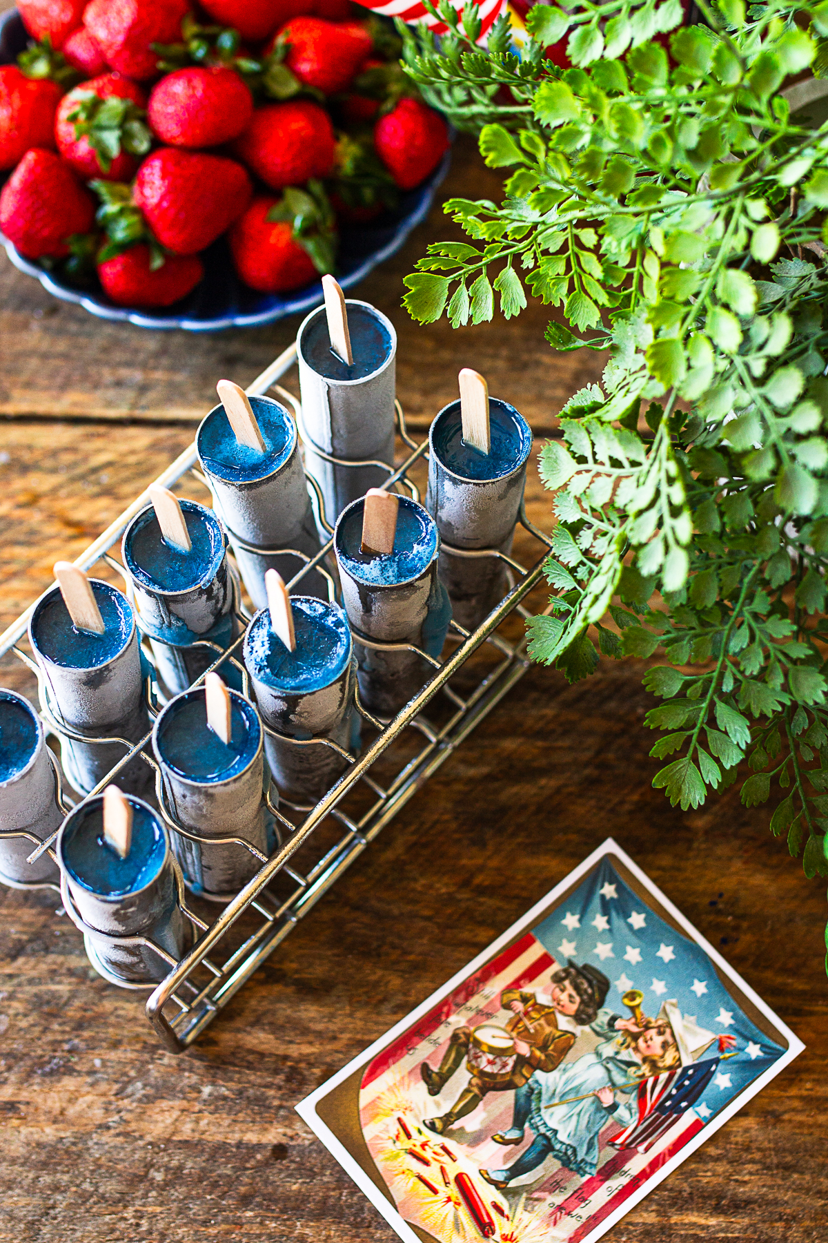 An overhead shot of frozen popsicles in a stainless steel rack showing only the vibrant blue top layer with wooden sticks inserted, sitting on a rustic wooden table next to fresh strawberries, green ferns, and a vintage 4th of July postcard.