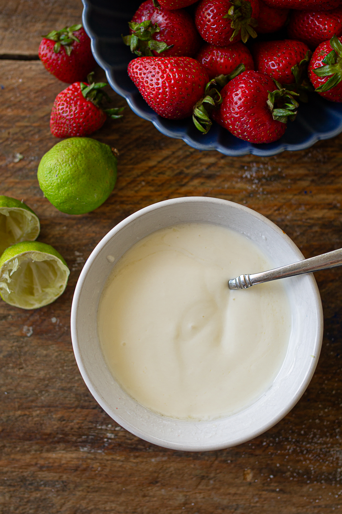 A white ceramic bowl filled with creamy vanilla yogurt and a spoon, sitting next to squeezed lime halves and fresh strawberries on a wooden table.