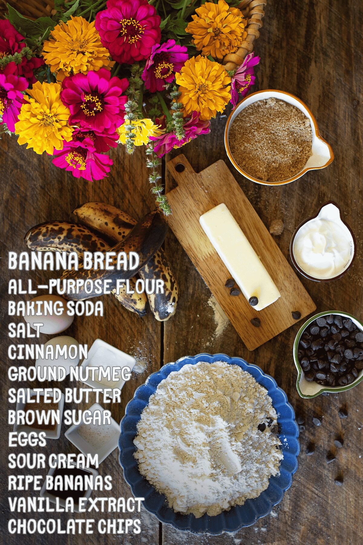 Flat lay of homemade banana bread ingredients including overripe bananas, flour, chocolate chips, butter, sour cream, brown sugar, and eggs on a rustic wooden table with fresh flowers.