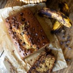 Overhead view of a freshly baked loaf of chocolate chip banana bread on parchment paper next to ripe spotty bananas and a bread knife.