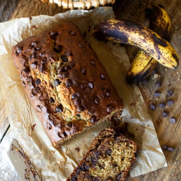 Overhead view of a freshly baked loaf of chocolate chip banana bread on parchment paper next to ripe spotty bananas and a bread knife.