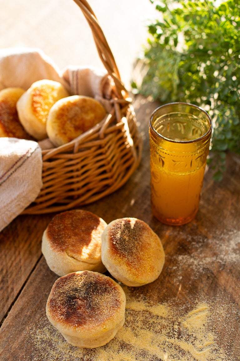 Golden homemade English muffins beside a wicker basket on a rustic wooden table.