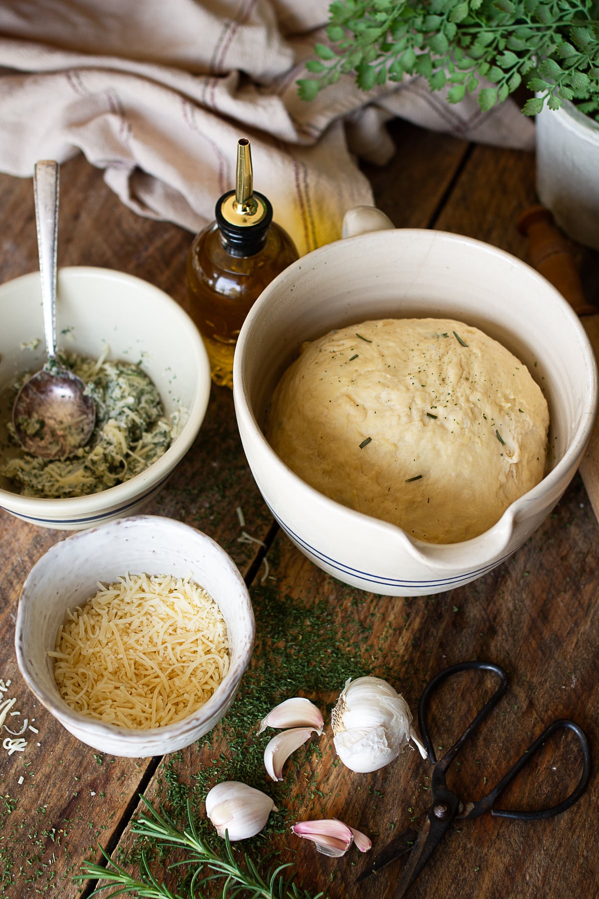 Homemade rosemary garlic bread dough in a mixing bowl surrounded by parmesan cheese, fresh garlic, rosemary, and herb butter.