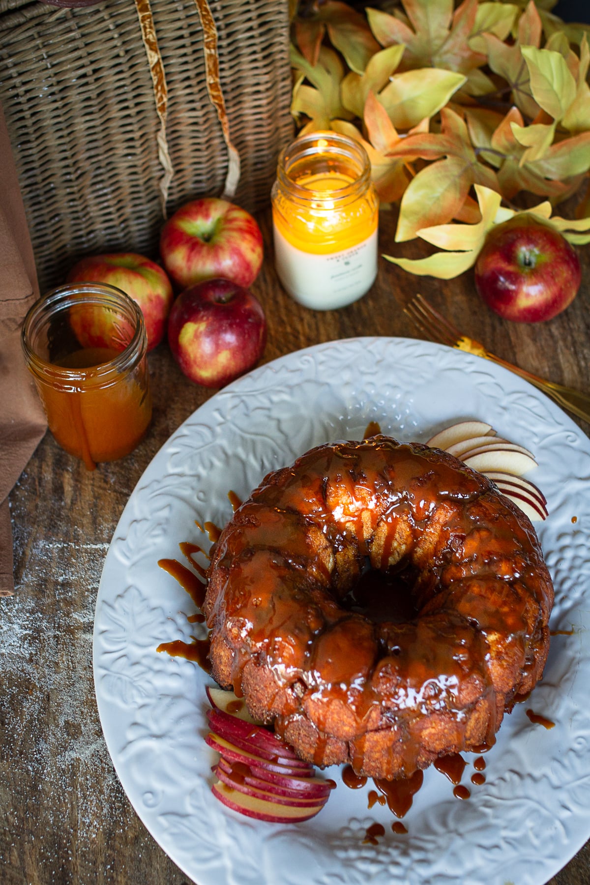 Overhead view of a golden caramel apple monkey bread ring served on a white platter next to a jar of caramel sauce and fresh red apples.