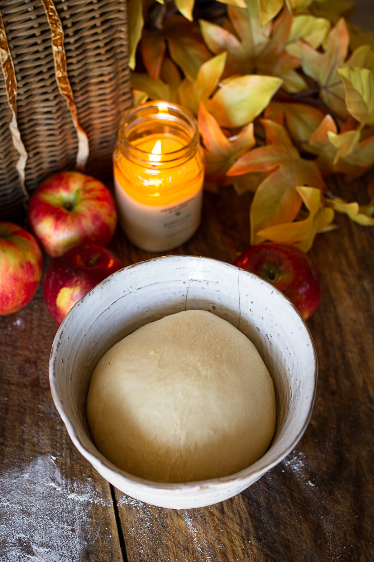 Smooth yeast bread dough rising in a white ceramic bowl on a wooden tabletop with fresh red apples in the background.